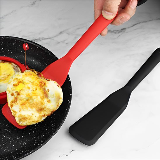 Un oeuf est sur une spatule rouge dans la poêle et a cote une spatule noir